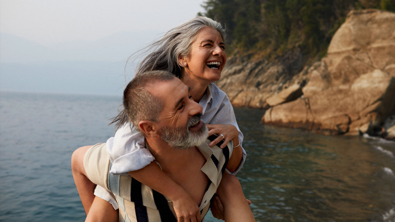 Couple &acirc;g&eacute; sur la plage. L&rsquo;homme, v&ecirc;tu d&rsquo;une chemise &agrave; manches courtes blanc cr&egrave;me &agrave; rayures longitudinales, porte la femme en blouse bleu clair sur son dos. Ils rient tous les deux.